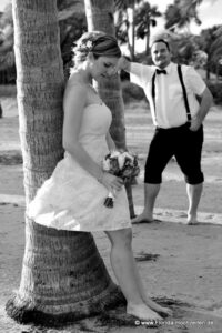 Braut und Braeutigam lehnen an Palmen sie schaut auf wunderschoenen Brautstrauss