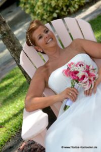 Glueckliche Braut im Adirondeck Beach Chair in weiss gekleidet mit Brautstrauss