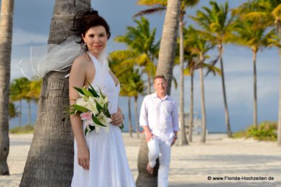 Miami Wedding auf Key Biscayne am Strand mit hunderten Palmen Braut lehnt an Palme und Braeutigam schaut im Hintergrund zu
