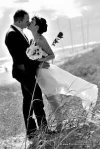 Hochzeitspaar in schwarz weiss mit Brautstrauss kuessend und Meer im Hintergrund und wehendem Brautkleid