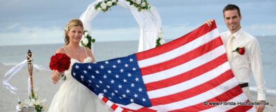Amerikanische Flagge wird von Brautpaar gehalten am Strand mit Rosenbogen in weiss geschmueckt mit Meer im Hintergrund